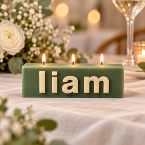 May include: A rectangular sage green candle with three lit wicks, featuring the name liam in raised, cream-coloured block letters on the front. The candle is positioned on a white fabric surface, with white flowers visible in the foreground and background.