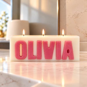 May include: A rectangular white wax candle with three lit wicks. The front of the candle features the name OLIVIA in large, raised, bright pink block letters. The candle is positioned on a smooth, light-coloured surface with a reflective finish.