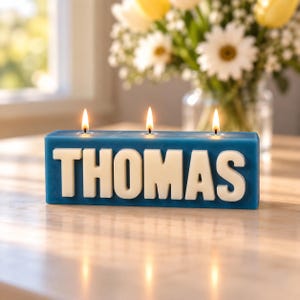 May include: A rectangular blue candle with three lit wicks, featuring the name THOMAS in large, raised white block letters on the front. The candle is positioned horizontally, with the wax surface showing three distinct flames burning brightly.