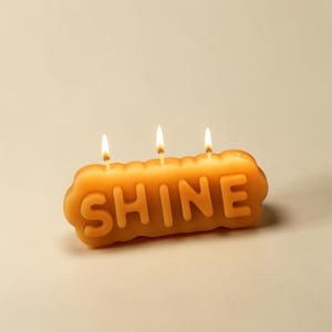 May include: A golden yellow wax candle shaped like a rounded rectangular block with the word SHINE embossed in raised capital letters on the front. The candle features three lit wicks positioned along the top edge, producing small, steady flames.