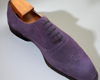 May include: A single purple suede wingtip shoe with black laces and a wooden shoe tree inside. The shoe has decorative perforations along the seams and a dark sole. The shoe is angled on a white surface.