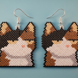 May include: A pair of beaded earrings shaped like a calico cat head. The design features a color palette of black, white, tan, and light brown beads arranged in a grid pattern. Each earring includes a silver-toned metal hook for attachment.