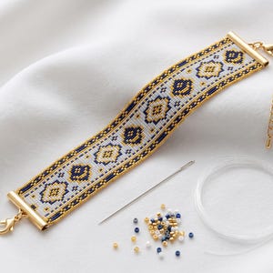 May include: A woven bead bracelet featuring a geometric pattern in white, navy blue, and gold, finished with gold-tone metal end bars and a lobster clasp. Included are a sewing needle, a coil of clear monofilament, and loose seed beads.