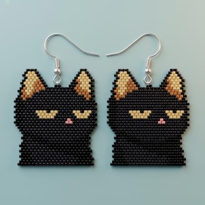May include: A pair of beaded earrings shaped like cat heads. Each earring features a black cat face with squinted yellow eyes, a small pink nose, and tan-colored inner ears. The earrings hang from silver-toned metal hooks for pierced ears.
