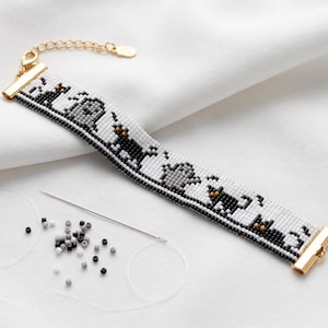 May include: A beaded bracelet featuring a repeating pattern of black cats, gray ghosts, and tombstones on a white background. The bracelet has gold-toned metal end caps and a lobster clasp. A sewing needle and loose black, white, and gray beads are nearby.