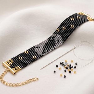 Puede incluir: Una pulsera de cuentas con fondo negro, una silueta de gato gris y pequeños detalles dorados. La pulsera tiene terminales de metal dorado y una cadena extensora. Se incluyen una aguja de coser, un hilo fino y cuentas sueltas negras y doradas.