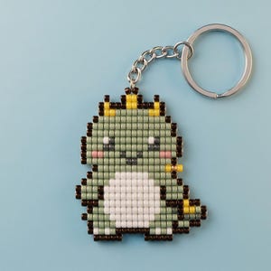 Peut inclure: Un porte-clés en perles représentant un petit dinosaure vert clair avec un ventre blanc, un contour marron foncé et une couronne jaune. La figurine est composée de petites perles carrées et est fixée à un anneau fendu en métal argenté.