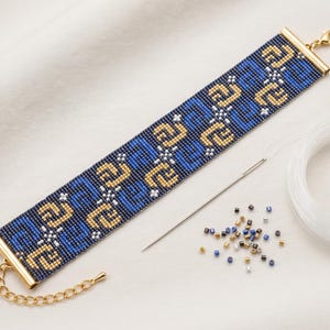 Peut inclure: Un large bracelet en perles avec un motif géométrique bleu marine, bleu roi et doré, agrémenté de petits accents blancs. Il possède des embouts en métal doré et une chaîne avec fermoir mousqueton. Une aiguille et des perles sont présentes.