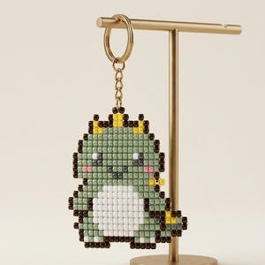 May include: A pixelated green dinosaur keychain with a gold-colored keyring and chain. The dinosaur has a white belly, pink cheeks, and yellow accents. The keychain is displayed on a gold-colored stand.