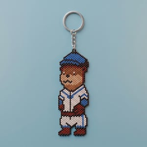 Könnte beinhalten: Ein Perlen-Schlüsselanhänger in Form eines braunen Bären, der eine blaue Baseballkappe und eine weiß-blaue Baseballuniform mit braunen Akzenten trägt. Die Figur besteht aus kleinen, gleichmäßigen Perlen und ist an einem silbernen Metallring befestigt.