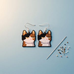 Op de afbeelding: Een paar bungelende oorbellen met een gepixeld ontwerp van een lapjeskat in zwart, bruin en wit. De kat heeft een sceptische uitdrukking met toegeknepen ogen. Zilverkleurige metalen haakjes zijn aan de bovenkant bevestigd. Een naald en kraaltjes liggen ernaast.