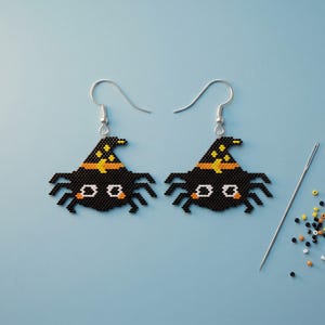 May include: A pair of beaded spider earrings featuring black bodies, orange hat bands, and yellow star accents on pointed hats, suspended from silver-toned metal hooks, accompanied by a single sewing needle and a small pile of loose seed beads.