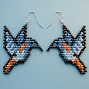 Peut inclure: Une paire de boucles d'oreilles pendantes formées de petites perles carrées représentant un oiseau. Le motif présente un contour noir avec des détails intérieurs bleu clair, blancs et orange. Chaque boucle est fixée à un crochet en métal argenté.