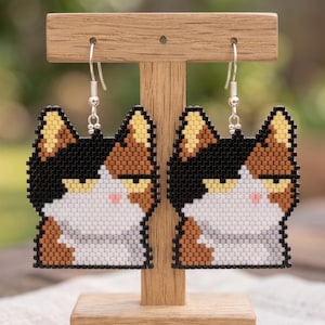 May include: A pair of cat-shaped earrings. The earrings are made of small, colorful beads in black, brown, white, and yellow. The cat's face has a grumpy expression. The earrings hang from silver-colored hooks and are displayed on a wooden stand.