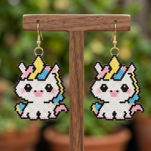 Puede incluir: Un par de pendientes de gancho dorados con diseños de unicornios pixelados. Los unicornios son blancos con detalles rosas, amarillos y azules. Los pendientes se muestran en un soporte de madera sobre un fondo verde borroso.