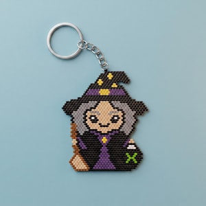 May include: A beaded keychain featuring a pixel-art style witch character. The witch wears a black hat with yellow stars and a purple band, a black robe with a purple center, and holds a brown broom. The keychain has a silver metal ring.