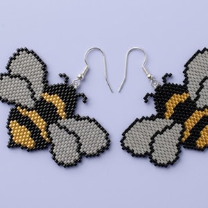 May include: A pair of dangle earrings crafted from small beads in a bee shape. The bees feature black, gold, and grey coloring with a pixelated appearance. Each earring is attached to a silver-toned metal hook for hanging from the earlobe.