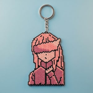 May include: A beaded keychain featuring a pixelated character design with pink hair, a pink visor, and a dark pink jacket. The figure is outlined in black beads and attached to a silver-colored metal split ring by a short metal chain.