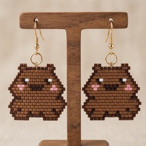 May include: A pair of handmade brown beaded earrings in the shape of a bear. The bear earrings have white eyes and pink cheeks. The earrings hang from gold-colored fish hook ear wires.