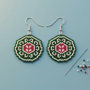 May include: A pair of octagonal beaded earrings featuring a geometric pattern in dark green, white, and magenta. The earrings hang from silver-toned metal hooks. A single sewing needle and several loose beads in matching colors lie nearby.