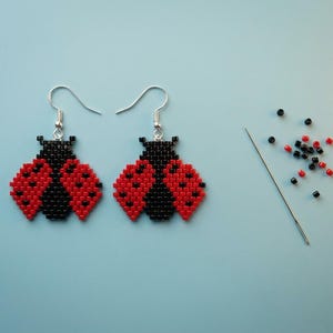May include: A pair of ladybug-shaped earrings crafted from small red and black square beads, featuring silver-toned metal hooks. Beside the earrings lies a thin metal sewing needle surrounded by a small collection of loose red and black beads.