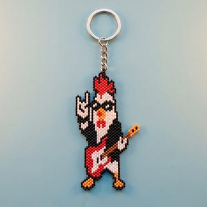 Pode incluir: Um porta-chaves de missangas com uma personagem de galinha em estilo pixel art, usando óculos de sol pretos e um casaco preto. A galinha faz um gesto de mão de rock and roll e toca uma guitarra elétrica vermelha e branca.