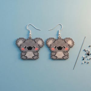 Könnte beinhalten: Ein Paar koalaförmige Ohrringe aus kleinen grauen, weißen, schwarzen und rosafarbenen Perlen. Jeder Ohrring hat einen silberfarbenen Metallhaken. Rechts daneben liegen eine einzelne Nähnadel und ein kleiner Haufen loser, passender Perlen.