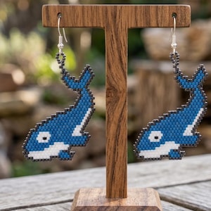 May include: A pair of handmade beaded earrings shaped like orcas. The earrings feature a pixelated design with blue, white, and black beads. They hang from silver-colored ear wires and are displayed on a wooden stand.