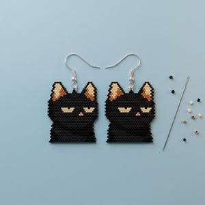 May include: A pair of beaded earrings shaped like black cats with tan inner ears and small pink noses, featuring silver-toned metal hooks. A single sewing needle and several loose, multi-colored seed beads are positioned to the right side.