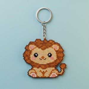 Op de afbeelding: Een kralen sleutelhanger in de vorm van een leeuw, gemaakt van kleine bruine, beige en roze kraaltjes. De leeuw heeft een grote manenbos, donkere ogen en een klein staartje, bevestigd aan een zilverkleurige metalen ring met een kort kettinkje.