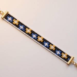 May include: A beaded bracelet featuring a geometric pattern of black, white, blue, and gold seed beads. The band is finished with gold-toned metal end caps and lobster claw clasps at both ends, creating a symmetrical and structured design.