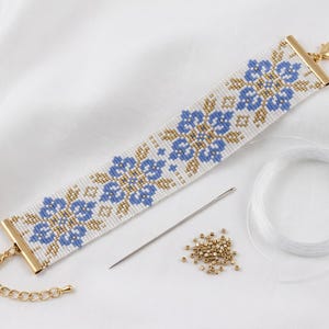 May include: A woven bead bracelet featuring a white background with a repeating floral pattern in blue and metallic gold. The bracelet has gold-tone metal end caps, a lobster clasp, and an extender chain. Included are a sewing needle, loose gold beads, and a spool of clear thread.