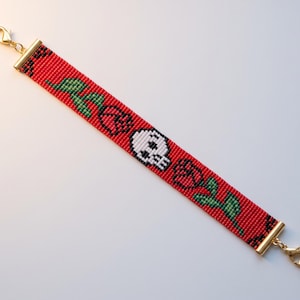 May include: A beaded bracelet featuring a red background with a pixelated white skull centered between two red roses with green stems and leaves. The bracelet is finished with gold-toned metal end bars and lobster claw clasps at both ends.