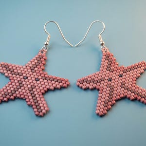 May include: A pair of starfish-shaped earrings crafted from small, square beads in shades of dusty rose and dark maroon. Each earring features a five-pointed star design with a central pattern of darker beads and includes a silver-toned metal hook.