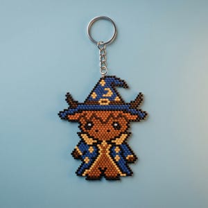 May include: A beaded keychain featuring a brown creature wearing a blue wizard hat and robe with yellow star and moon accents. The design is crafted from small square beads in a pixelated style, attached to a silver metal split ring keychain.