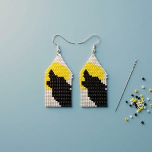 May include: A pair of beaded drop earrings featuring a geometric design of a black wolf silhouette against a yellow moon and white background. The earrings have silver-toned metal hooks and are displayed with a sewing needle and loose beads.
