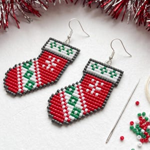May include: A pair of beaded earrings shaped like Christmas stockings, crafted with red, white, green, and dark gray seed beads. The design features a geometric pattern on the cuff and a diagonal stripe motif on the foot, with silver-tone hooks.
