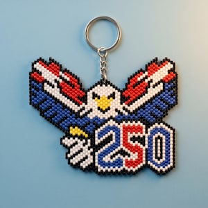 May include: A beaded keychain featuring a pixelated bald eagle with outstretched wings in red, white, and blue. The eagle has a yellow beak and white head. Below the eagle, the number 250 is displayed in red, white, and blue beads.