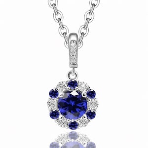 Op de afbeelding: Een zilverkleurige ketting met een ronde hanger met een grote, ronde, diepblauwe steen in het midden. De centrale steen is omringd door een halo van afwisselend kleinere ronde blauwe stenen en heldere witte stenen, met extra stenen op het oogje.
