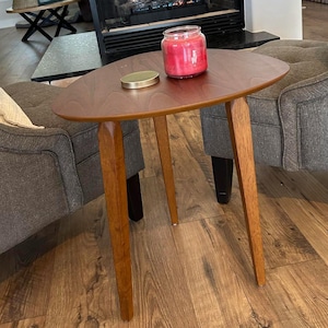 May include: A mid-century modern style oval side table with three wooden legs. A red candle in a glass jar and a gold lid sit on the table. The table is placed between two gray upholstered chairs.