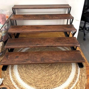 May include: A tiered display stand with five shelves. The shelves are made of dark brown wood and supported by black metal frames. The stand is placed on a round, woven mat with a white border, suitable for displaying products.