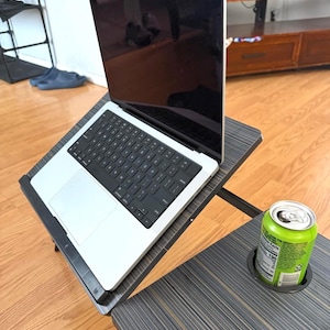 May include: A silver laptop with a black keyboard sits on a black and gray adjustable laptop desk. A green can of soda rests in the built-in cup holder of the desk. The desk is on a wooden floor.