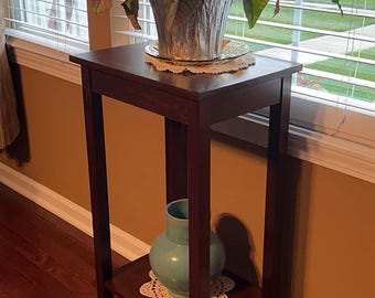 May include: A dark brown wooden plant stand with a small shelf. A potted plant with green leaves and red accents sits on top. A light blue vase rests on the lower shelf, with a decorative doily under each item.