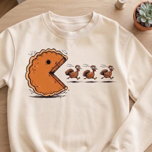 May include: A cream-colored crewneck sweatshirt featuring a graphic print of a large orange pumpkin pie slice shaped like a video game character chasing three brown turkeys that are running away in a line across the front of the garment.