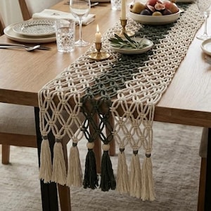Puede incluir: Un camino de mesa de macramé largo con un intrincado patrón de nudos en cordón de algodón color crema y verde oscuro. El camino está rematado con borlas decorativas y cuentas de madera en ambos extremos, sobre una mesa de comedor de madera.