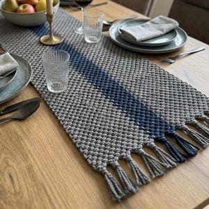 May include: A wooden dining table set for a meal. A gray and blue woven table runner with tassels runs down the center. Plates, silverware, glasses, candles, and a bowl of fruit are arranged on the table.
