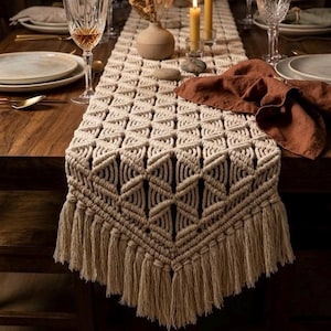 Puede incluir: Una mesa de comedor de madera con un camino de mesa de macramé beige con flecos, platos, vasos, velas y un jarrón con flores secas. La mesa está preparada para una comida, creando un ambiente cálido y acogedor.