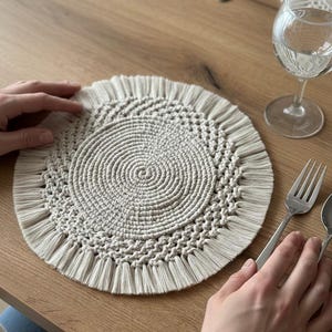 May include: A beige macrame placemat with a spiral design and fringe detail. A glass of water, a fork, and a spoon are on a wooden table. Another placemat and a knife are also visible.