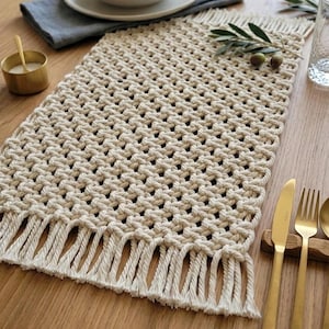 May include: A cream macrame table runner with a fringed edge, on a wooden table. The rectangular runner has a woven pattern. The table setting includes a white plate, bowl, and gold cutlery. The runner is approx. 40 cm wide.
