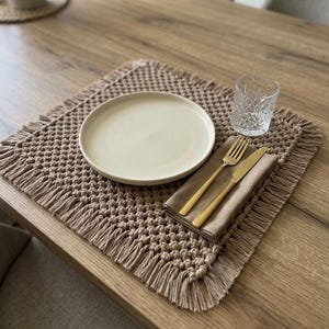 May include: A square, beige macrame placemat with a knotted texture and fringed edges. On the placemat sits a round, off-white ceramic plate, a folded tan cloth napkin, a gold-toned fork and knife, and a clear, textured glass tumbler.